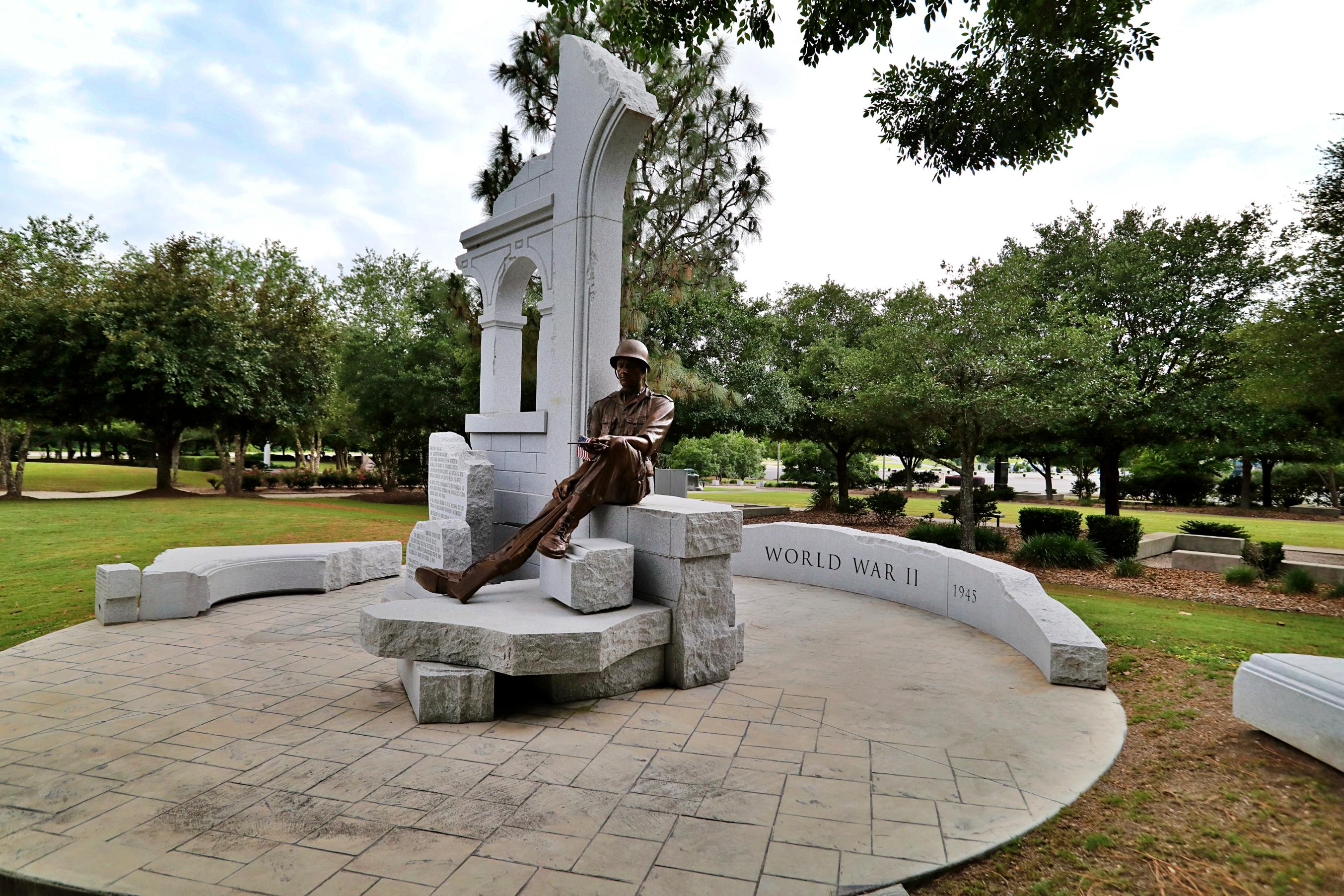A statue of a person sitting on a stone bench in a park.